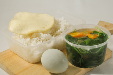 a menu of white rice with clear vegetables of spinach leaves and carrots, side dishes of shrimp crackers and a salted egg on a wooden board isolated on a white background