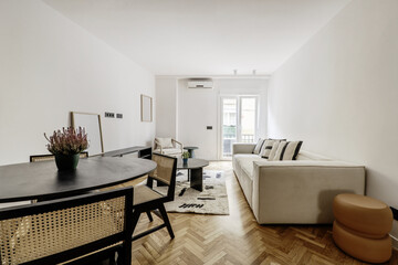 Living room of a modern home with a black circular wooden table, matching chairs with slatted backs and seats, a long sofa upholstered in beige fabric and a balcony in the background