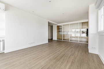 Large empty modern living room with white painted walls and sliding glass partition enclosing a kitchen with wooden furniture