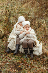 Two twin sisters are sitting in an autumn park, wrapped in a white blanket, relaxing.