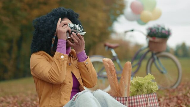 Cute black woman spending leisure time in park, sitting on yellow blanket of fallen leaves, taking pictures on vintage camera. Hobby. Cycling, shopping. High quality 4k footage