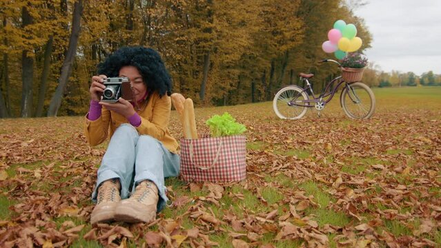 Colored female spending leisure time capturing bright moments on vintage camera sitting on lawn blanketed with yellow fallen leaves. Relax after shopping. High quality 4k footage