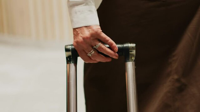 Older woman carries trolley bags at hotel reception, approaching front desk to do check in and start vacation trip. Traveller with luggage entering lobby, senior travel. Handheld shot. Close up.