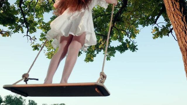 Child plays on wooden swing, dreams of flying. Happy little girl swings on swing in park under tree at sunset. Baby swing, kid girl smile in flight. Concept of family happiness, dreams, entertainment