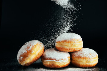 Powdered sugar is poured onto donuts. Traditional Jewish dessert Sufganiyot on black background. Cooking fried Berliners. - obrazy, fototapety, plakaty