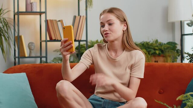Young blonde woman blogger taking selfie on smartphone, communicating video call online with subscribers, recording stories for social media. Adult girl at home apartment living room sitting on couch