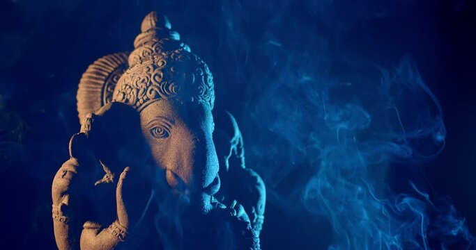 Ganesha idol made of sandstone on a black background at night. There is warm light and blue light. There is smoke from the incense that people burn to worship.