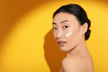 Beautiful young woman in sunlight with sun protection cream on her face against orange background,...