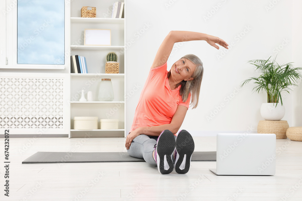 Sticker Senior woman in sportswear stretching near laptop on mat at home