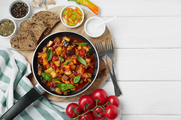 Frying pan with tasty ratatouille, ingredients and bread on white wooden table, flat lay. Space for...