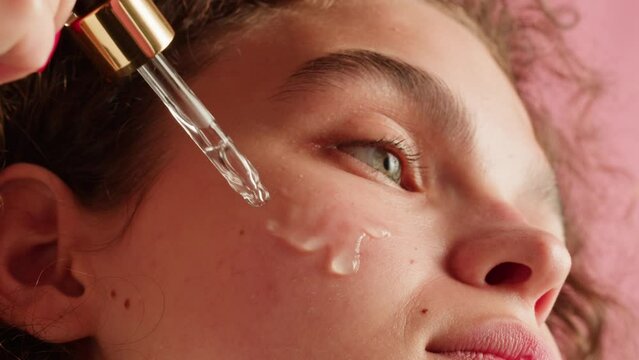 Young woman applying hydrogelic serum cream on her face close-up. Morning skin care routine. Pink background