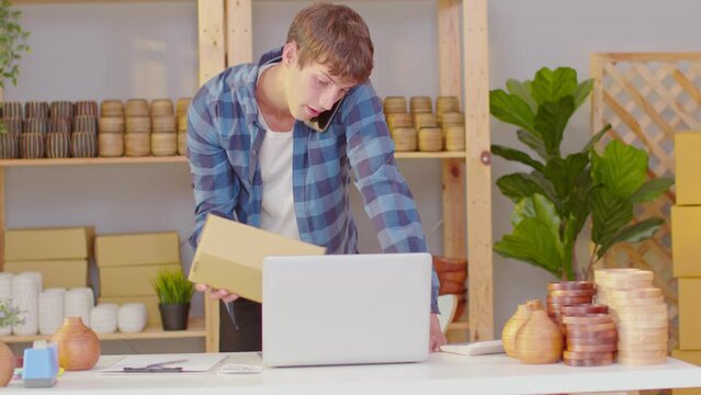 Happy Caucasian young man business owner flower vases store working and selling online with computer laptop smile and talking with customer to get order in mobile phone. Small business concept