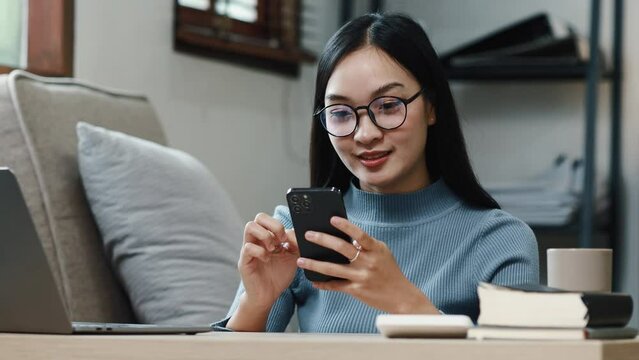 An Asian woman is playing with a smartphone in the living room at home. She spends her free time on her weekend break. She is a company employee. Long vacation. Long holiday vacation.