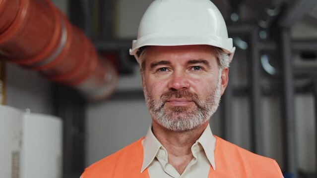Portrait of factory employee wearing white hard and orange vest hat posing at workplace. Civil engineer with grey beard and hair making eye contact after doing his complicated job at technical room.
