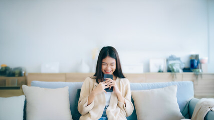 Happy young asian woman relax on comfortable couch at home texting messaging on smartphone, smiling...