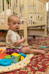 The baby is sitting on the floor near the crib and playing with toys.