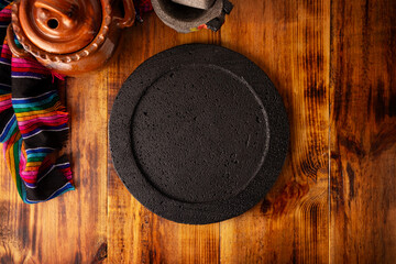 Empty stone comal plate on wooden rustic table, colorful typical mexican fabric with clay pot and stone molcajete, traditional kitchen utensil used in Mexican gastronomy.