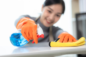 Asian woman wearing protective gloves smiling and wiping dust using spray and rag while cleaning house in close-up