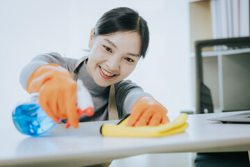 Asian woman wearing protective gloves smiling and wiping dust using spray and rag while cleaning house in close-up