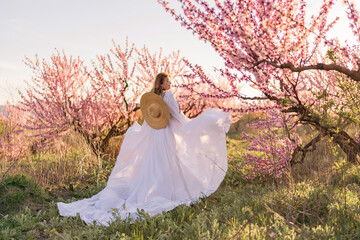 Woman blooming peach orchard. Against the backdrop of a picturesque peach orchard, a woman in a long white dress and hat enjoys a peaceful walk in the park, surrounded by the beauty of nature.