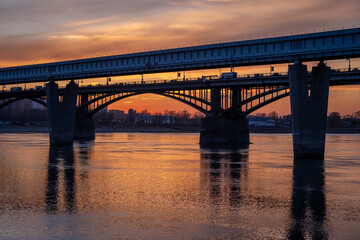 Fototapeta na wymiar Beautiful view of the bridge over which cars drive at sunset. A river flows under the bridge, reflecting the sunset rays. Bridge in the city of Novosibirsk, Russia