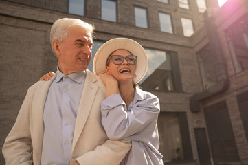 A woman in a hat and glasses hugs an elderly man in a white jacket from behind. Romantic relationships of mature people. 