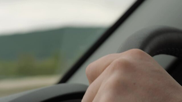 Man hand holds steering wheel driving car along rural road against mountains