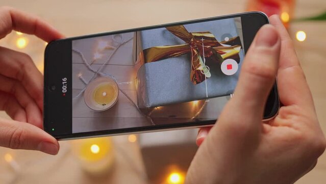 Unidentified woman recording video on her smartphone, capturing festively decorated table with candlelight and New year gift box. Festive Christmas wrapping.