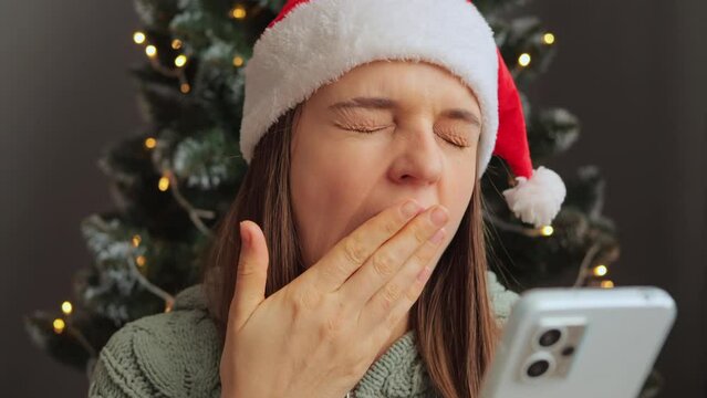 Yawning woman wearing Santa Claus hat using phone near Christmas tree in the decorative interior feeling fatigue from online scrolling feeling sleepy.