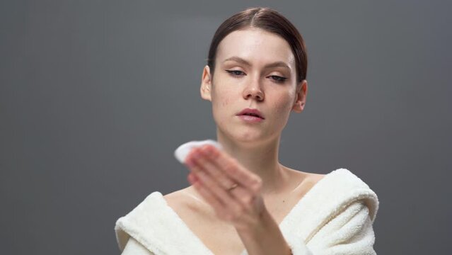 Cute woman removing makeup with cotton pad and micellar water. Girl with perfect skin on gray background. Beauty and skin care concept