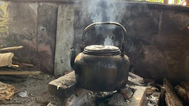 Old black kettle stands on fire made of firewood in the old fashioned kitchen. Kettle over the fire. An old vintage smoked teapot is boiling water