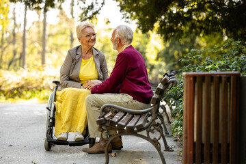 Senior man and his wife in wheelchair talking in enjoying in the park. - obrazy, fototapety, plakaty