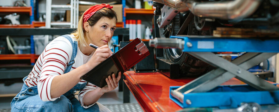 Concentrated mechanic woman with clipboard reviewing damages in motorbike over platform on workshop