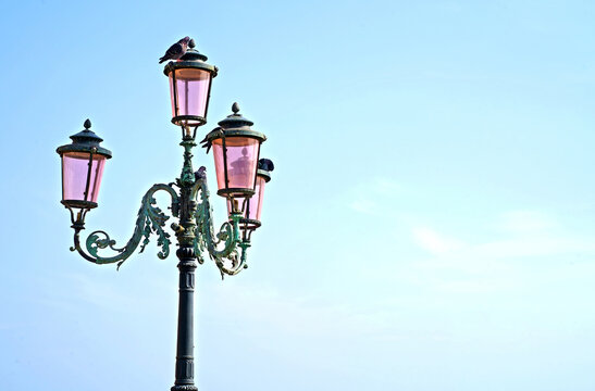 Lantern on the street of Venice. The famous pink lights of Venice. Four lamps with pink glass are on an ornate black metal lamp post. Blue cloudless sky.