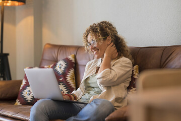 One woman doing video call online at home sitting on sofa using laptop on her legs. Relax and online activity. Modern female adult people using computer and smile touching her neck shy and happy