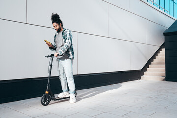 Trendy smiling bearded man in casual clothes riding electric scooter in urban background. Handsome...