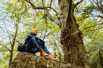 View from below of a handsome man dressed with winter outerwear clothes, looking up sitting in the forest in front of a big tree.