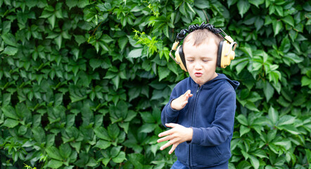 A positive little boy of 6 years old in yellow retro headphones listens to music and dances. A boy dances against a wall of plants. Banner with space for text