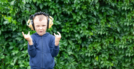 A positive little boy of 6 years old in yellow retro headphones listens to music and dances. A boy dances against a wall of plants. Banner with space for text