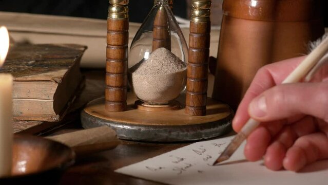 Writing on paper with a goose feather quill pen and ink by candlelight – An antique hourglass and book in the background