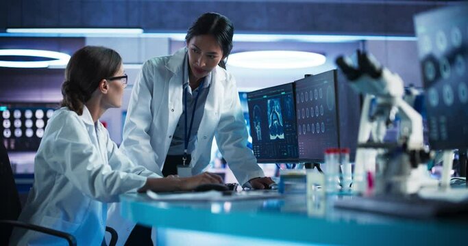 Medical Hospital Research Laboratory: Caucasian And Asian Female Scientists Talking And Using Desktop Computer With MRI Scans Of Human Brain. Surgeon And Neurologist Discussing Brain Damage Treatment.