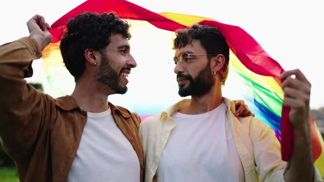 Intimate moment under rainbow flag, gay men share loving gaze, symbolizing LGBTQ+ pride and joy of being true to oneself. Male couple smile reflecting world reflecting world where love knows no bounds