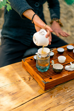 The hands of a professional tea master who pours fresh natural green tea from a glass teapot into bowls on wooden chaban table