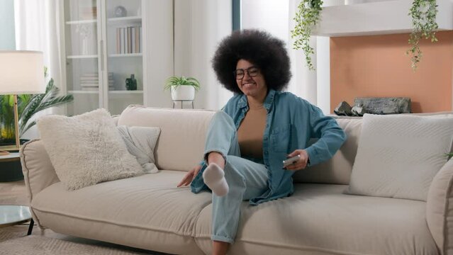 African American girl ethnic woman female at home walk come in living room sit on couch looking mobile phone app put smartphone away relax resting on comfortable sofa relaxing smiling weekend peace