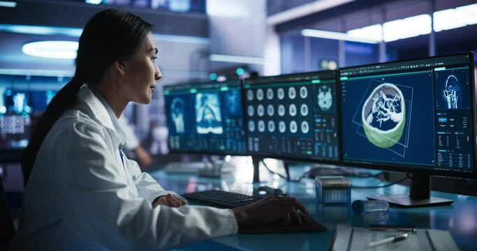 Female Asian Neurologist Using Desktop Computer With Human Brain MRI Scans In Modern Medical Research Center Laboratory. Professional Doctor Looking At Patient's Brain In 3D Software, Finding Tumors.