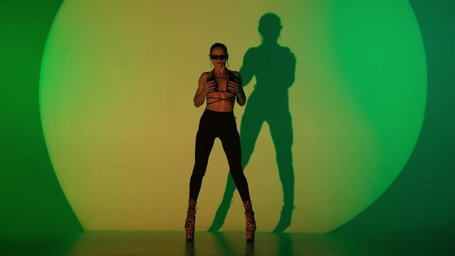 Appealing woman dancing modern choreography in leather top on high heels in the studio in green spotlight neon background.