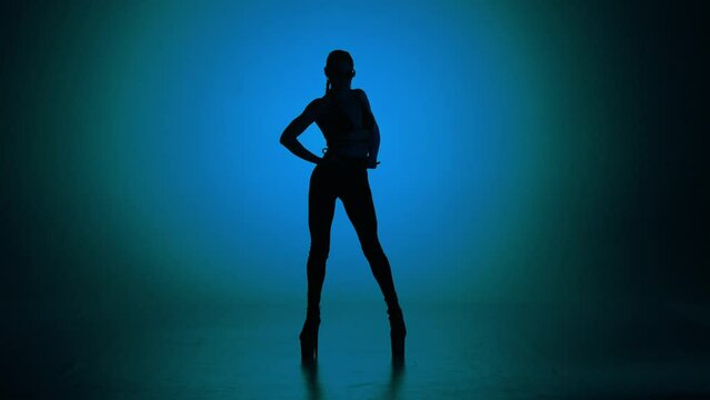 Silhouette of appealing woman dancing modern choreography in leather top on high heels in the studio on blue neon background.