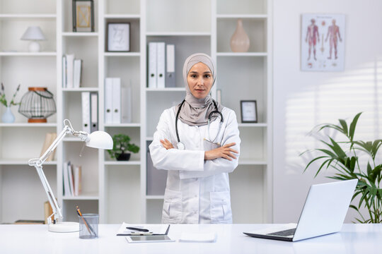 Portrait of serious confident successful female doctor in hijab in white medical coat looking at camera with crossed arms, clinic worker working inside office medical room.
