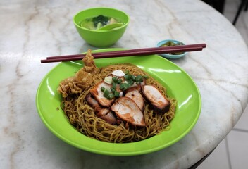 Dry noodles in sauce with toppings of fork roasted pork, green onions and fried wonton. The dish is accompanied by a bowl of soup. Common street food in Malaysia known as wonton noodles. 