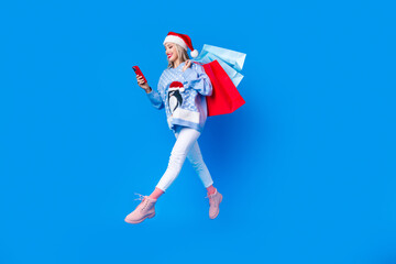 Full body cadre of browsing smartphone young blogging woman run jump air wear red santa hat bring bags isolated on blue color background